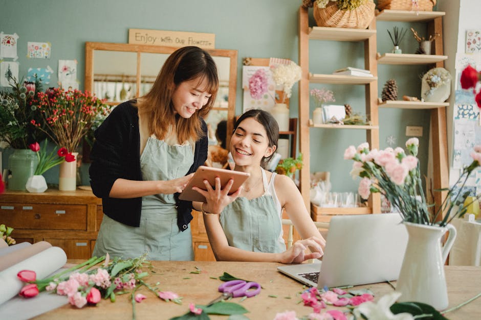 Two women in a florist shop look at a tablet while surrounded by various fresh flowers, including pink carnations, red tulips, and other floral arrangements. One woman with shoulder-length brown hair 