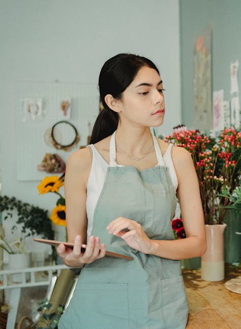 A young woman with long dark hair, dressed in a white sleeveless top and a light green apron, stands in a floral shop holding a smartphone in her left hand. Behind her, an assortment of fresh flowers,