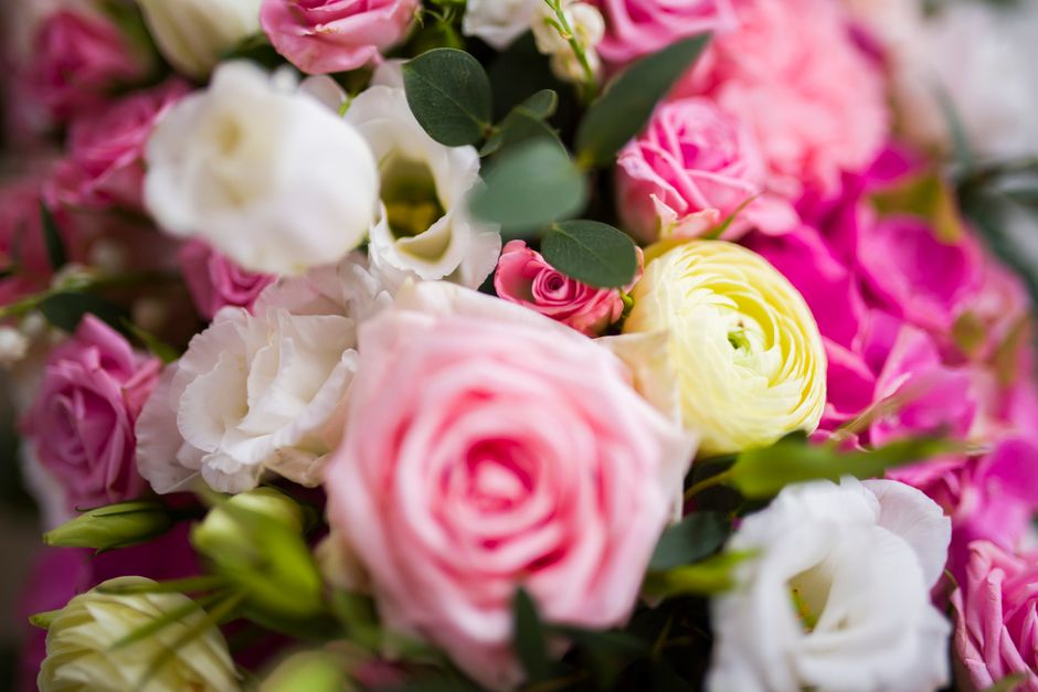 A close-up view of a vibrant floral bouquet featuring soft pink roses, creamy white lisianthus, and delicate yellow ranunculus, accented with dark green eucalyptus leaves. The flowers are arranged in 