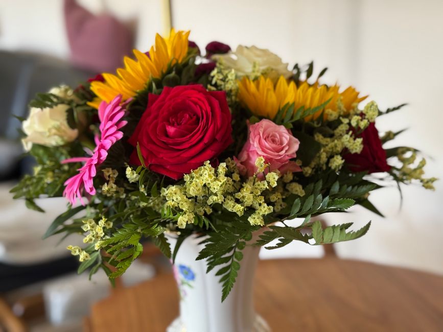 A vibrant floral bouquet arranged in a white ceramic vase with a delicate floral design. The arrangement features a dominant red rose at the center, surrounded by soft pink roses, bright yellow sunflo