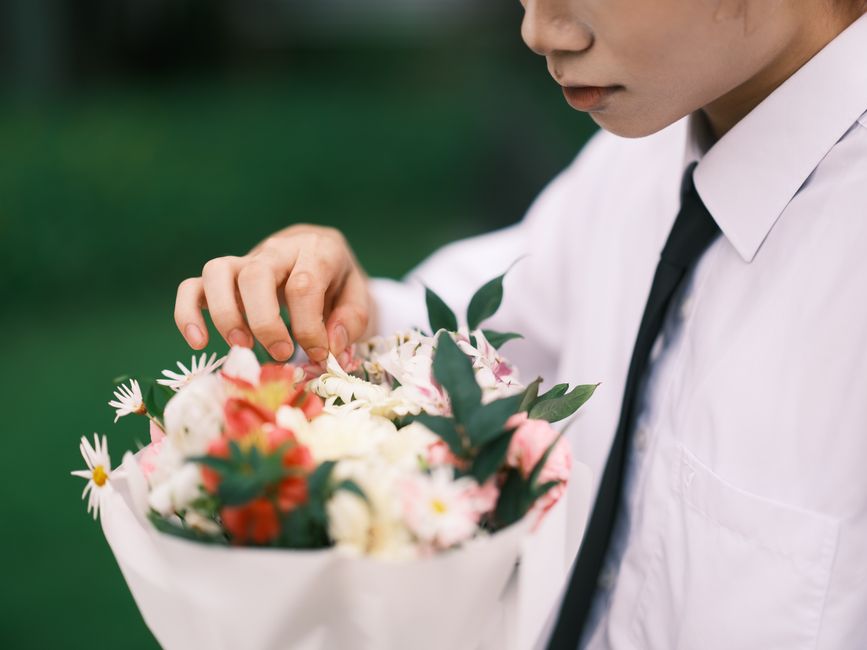 A person dressed in a white shirt and black tie is holding a round bouquet arrangement of fresh flowers. The bouquet features a mix of pink, white, and red blossoms, including roses, daisies, and smal
