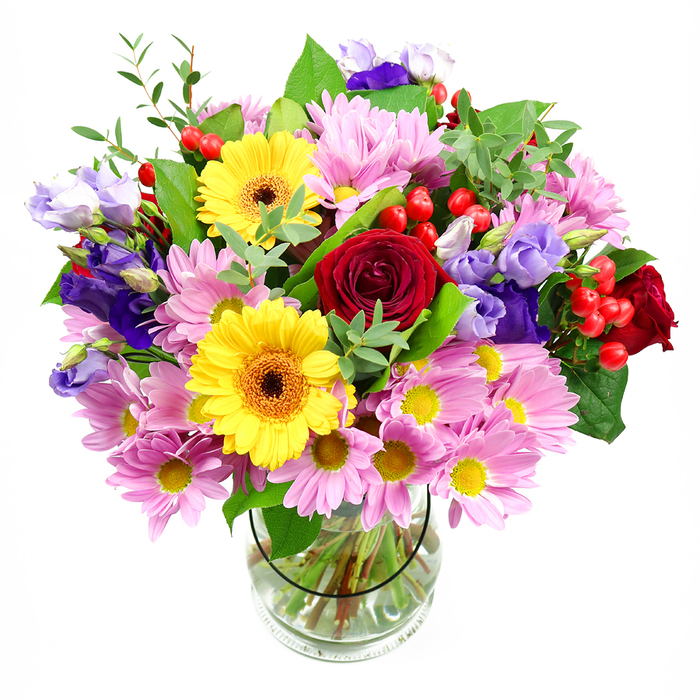 A clear glass vase holds a sumptuous bouquet of mixed flowers, its colourful dome shape set against a clean white background that allows every petal and leaf to stand out. At the centre of the design, rich red roses blossom in velvety, layered spirals, providing an elegant focal point and a touch of classic romance. Just alongside them, two bright yellow gerbera daisies, each with a dark brown-orange centre, burst forth like small suns, bringing a sense of joy and energy that immediately catches the eye. These bold flowers are gently framed by plentiful soft pink daisies or chrysanthemums, their numerous slender petals encircling tiny yellow centres and creating a light, cloud-like texture around the heart of the bouquet. Interwoven throughout are stately purple and lavender lisianthus flowers, their bell-shaped blooms and slightly ruffled edges lending a cool, graceful contrast and a sense of gentle movement. Dotted carefully between petals, clusters of glossy red hypericum berries add small, rounded shapes and a lively textural accent, making the bouquet feel modern and thoughtfully detailed. The flowers rest within a nest of lush greenery: broad, dark leaves offer a solid base, while more delicate, light-green sprigs extend outward, hinting at the kind of foliage you might notice in well-tended Cheam gardens or along local residential streets near Cheam station. Through the transparent vase, you can see the neatly arranged stems submerged in clear water, emphasizing the freshness and professional care behind the arrangement. This bouquet feels ideal for celebrations in Cheam-from birthday lunches at home to quiet anniversaries-bringing together vibrancy, softness, and a welcoming, homely charm.